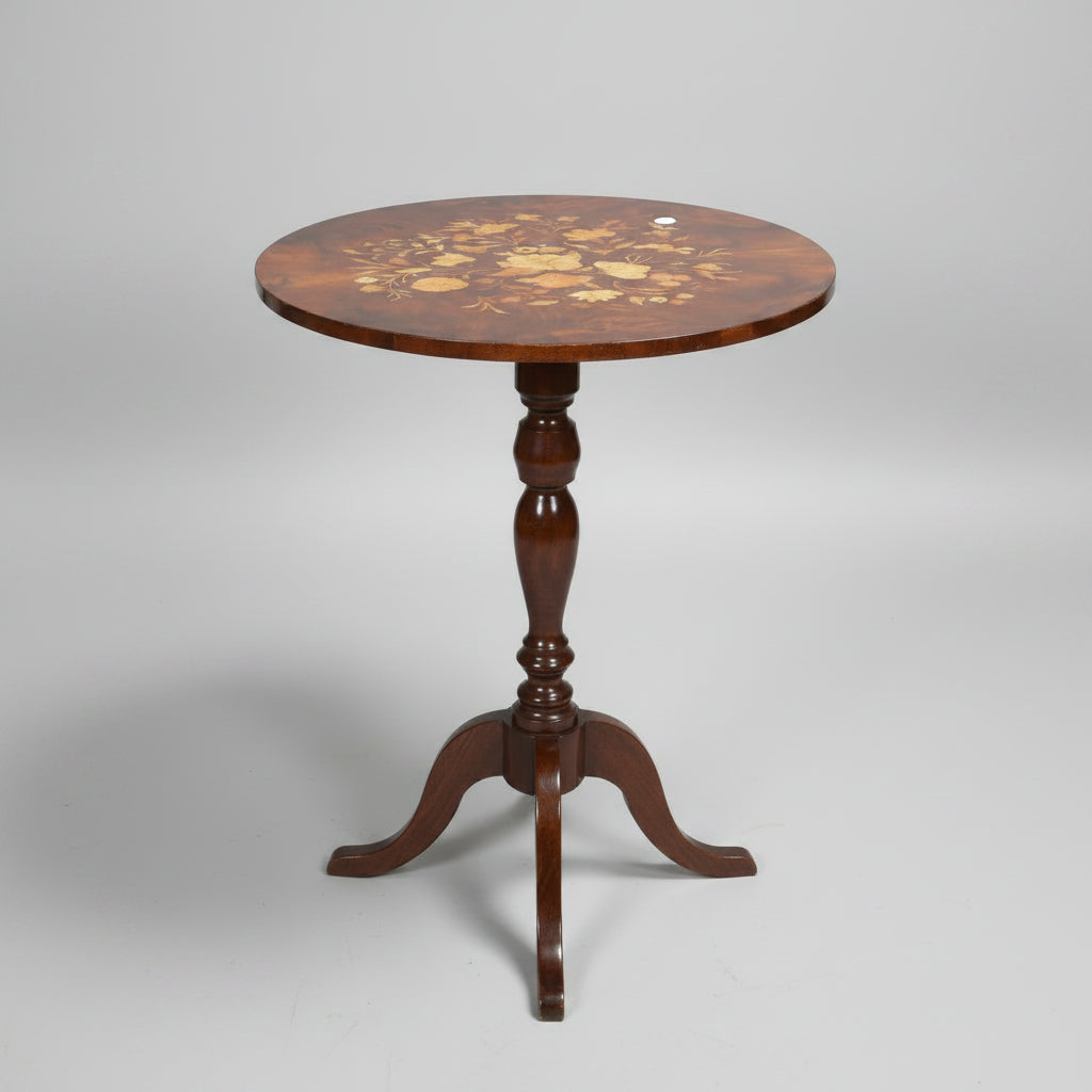 A round wooden pedestal table with an inlaid floral pattern on the tabletop, surrounded by other furniture in what appears to be a shop or storage area.