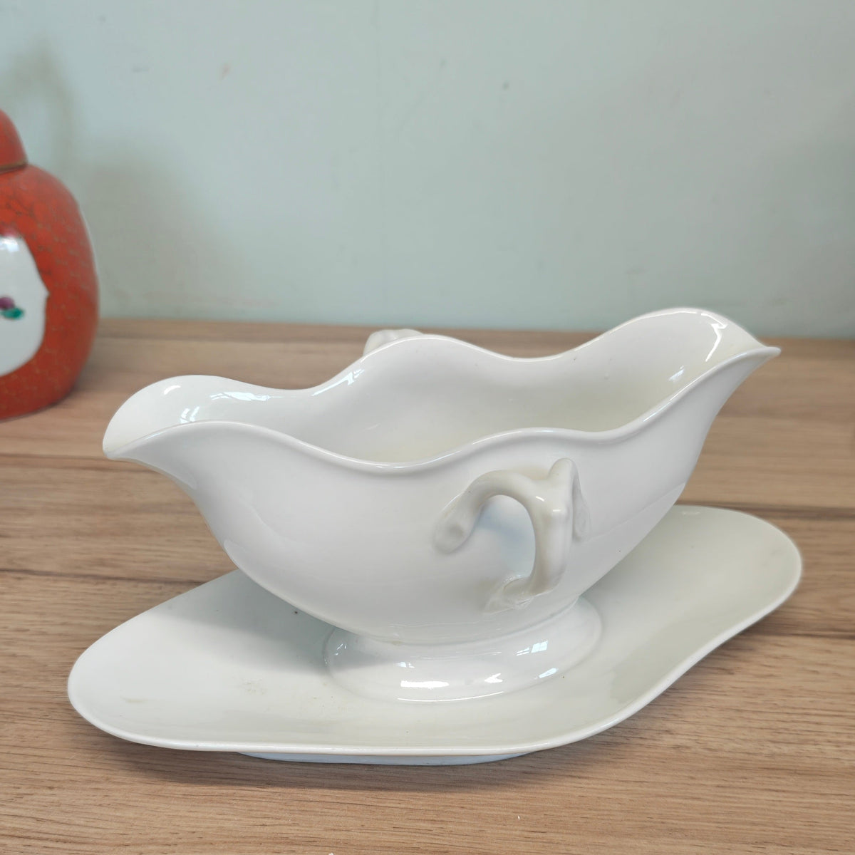 A white ceramic gravy boat with a matching saucer, placed on a light wood tabletop against a pale wall.