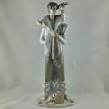 A glazed ceramic figurine of a young boy holding a goat on his shoulder, standing on a base with a dog at his feet, in a light gray and white glaze against a neutral gray background.