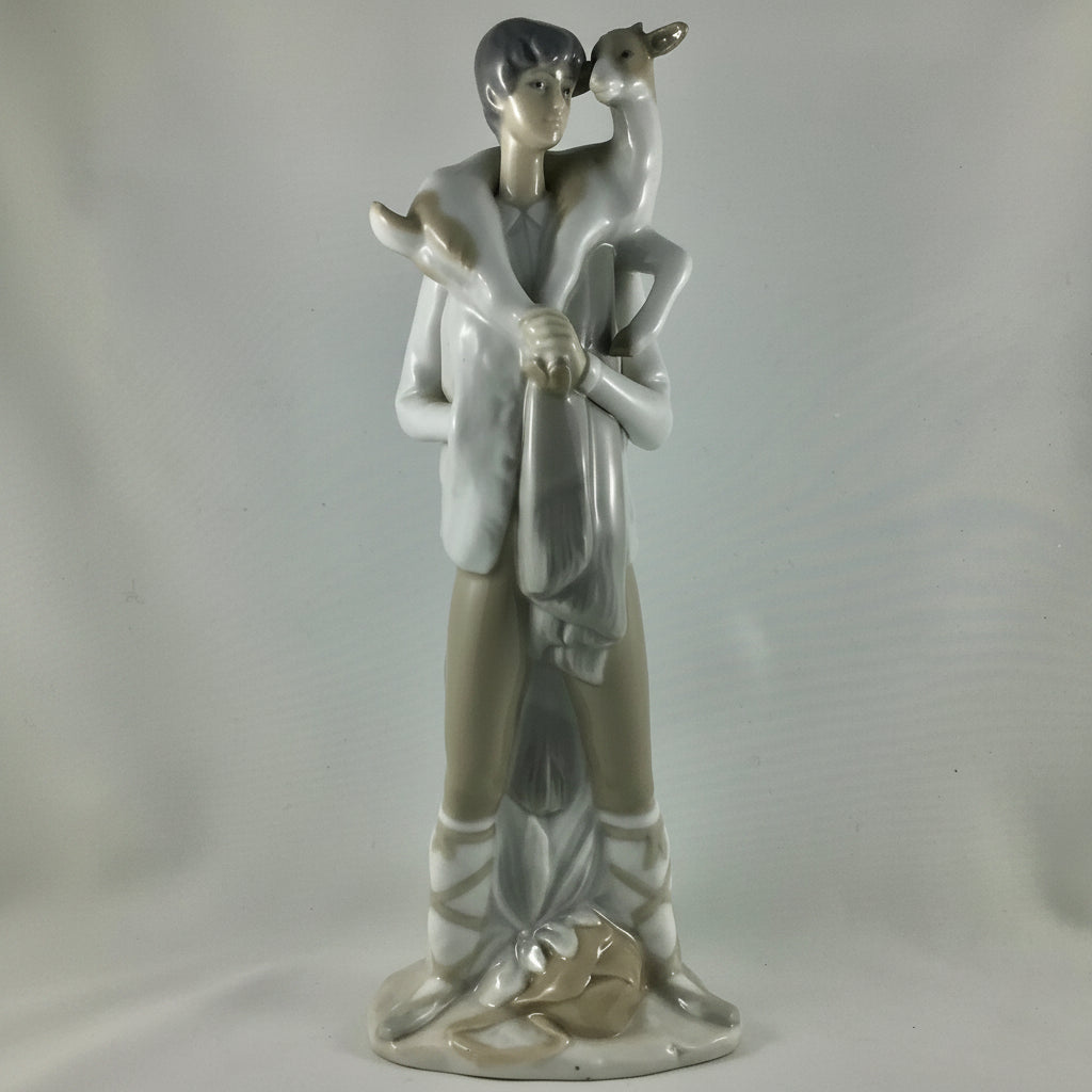A glazed ceramic figurine of a young boy holding a goat on his shoulder, standing on a base with a dog at his feet, in a light gray and white glaze against a neutral gray background.
