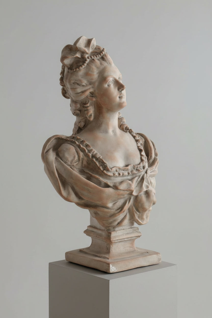 A classical-style stone bust sculpture of a woman with ornate hair and draped fabric, displayed on a pedestal in an indoor setting with light-colored paneled walls and dried pampas grass.