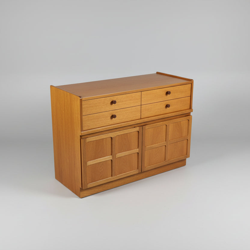 A mid-century teak compact sideboard or cupboard with four drawers and two cupboards, placed against a white wall on a wooden floor.