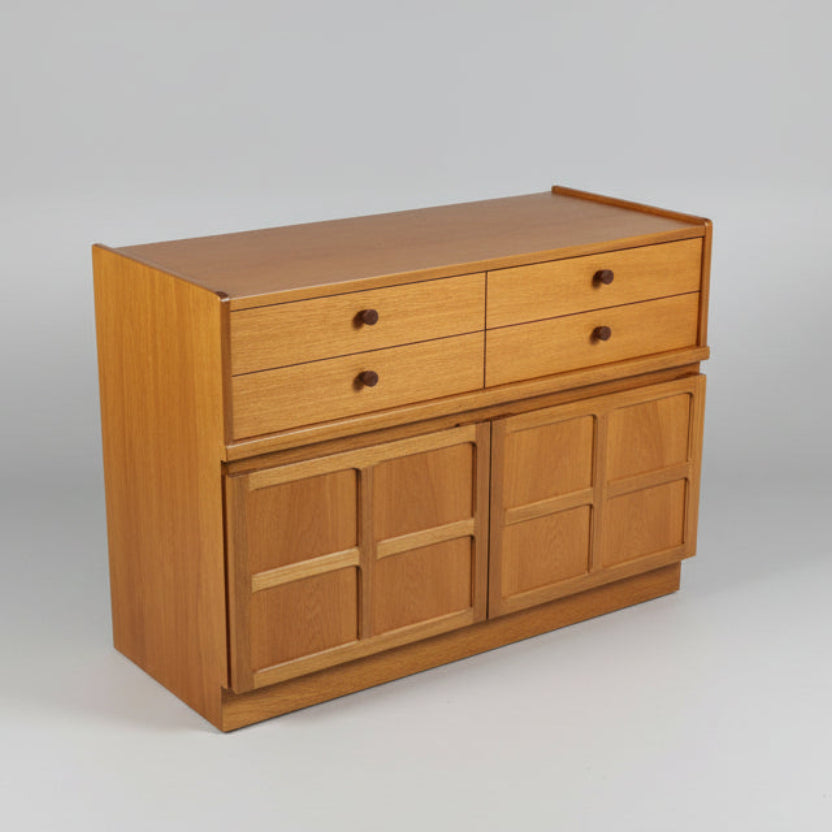 A mid-century teak compact sideboard or cupboard with four drawers and two cupboards, placed against a white wall on a wooden floor.
