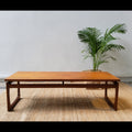 A brown, mid-century style floating top coffee table with a simple, curved design, displayed in a room with a white wall and a green potted plant in the background.