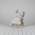 A porcelain figurine of a young girl in a white dress leaning against a tree with birds and swans, shown on a plain white background.