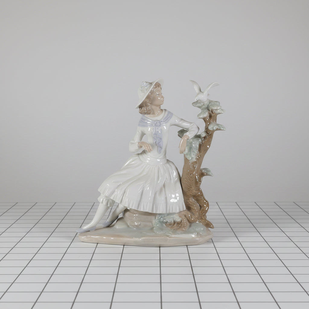 A porcelain figurine of a young girl in a white dress leaning against a tree with birds and swans, shown on a plain white background.