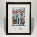 Framed sports memorabilia featuring a printed Manchester City Champions League trophy celebration, mounted with a white mat and black frame, with a small metal plaque below.