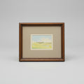 A framed landscape art print featuring a yellow-green field under a pale blue sky, set in a brown wooden frame with a gray mat on a wooden surface.