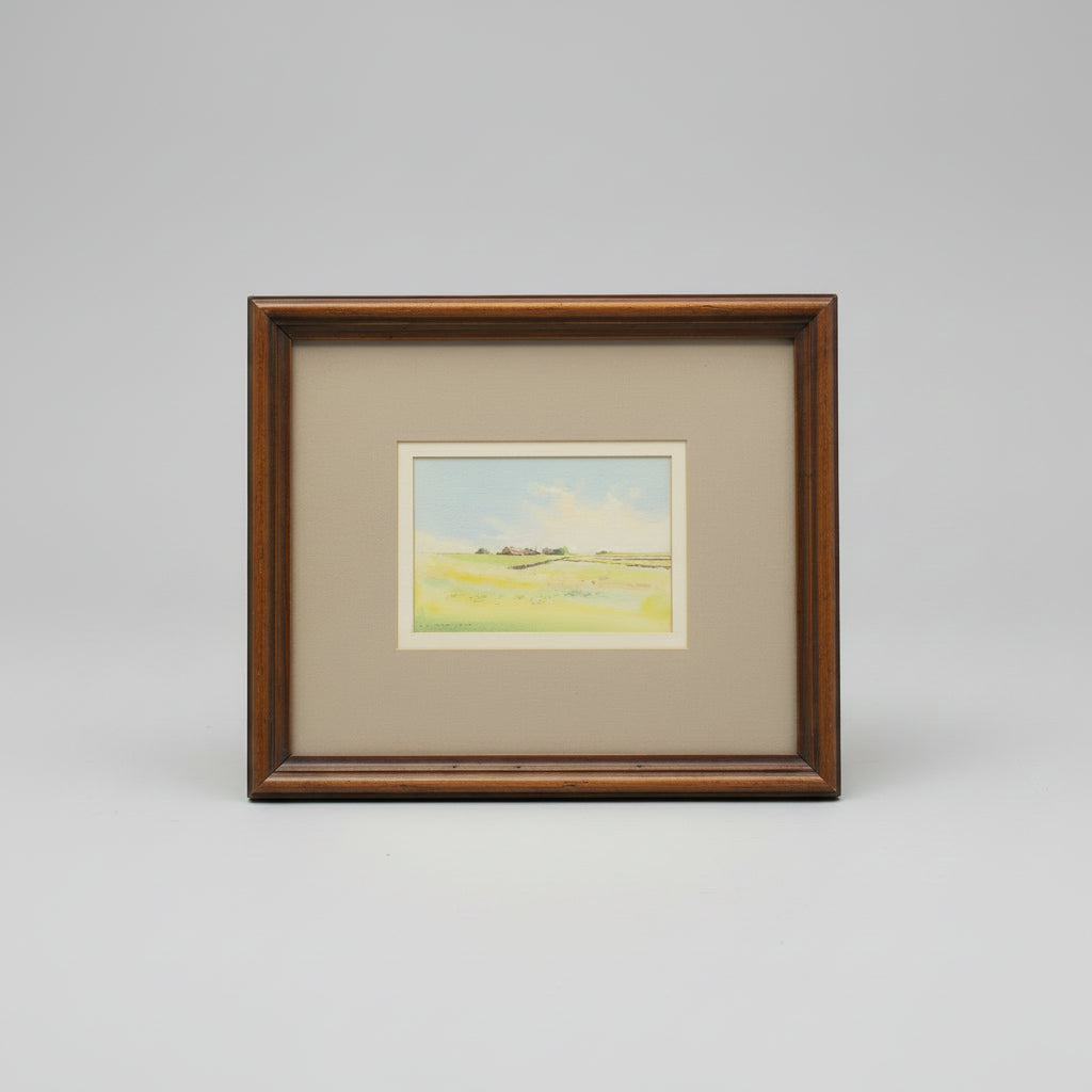A framed landscape art print featuring a yellow-green field under a pale blue sky, set in a brown wooden frame with a gray mat on a wooden surface.