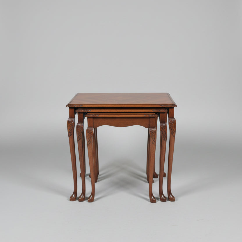 A set of nested wooden nesting tables with a glossy marquetry top and carved legs, displayed in a furniture showroom alongside a red leather armchair and a wooden chest of drawers.