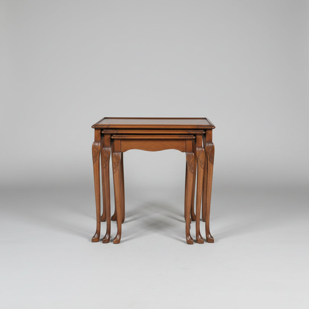A set of nested wooden nesting tables with carved cabriole-style legs, displayed in a furniture showroom alongside other vintage pieces including a leather armchair and a wooden chest of drawers.