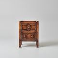 Wooden side table with two drawers on a red patterned rug, with a painting in the background.
