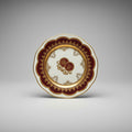 A decorative porcelain plate with gold, brown, and white colors, featuring floral patterns and a central motif, displayed on a lace doily.