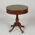 A round wooden pedestal table with a dark green and gold patterned top, photographed in a cluttered antique shop or furniture store.