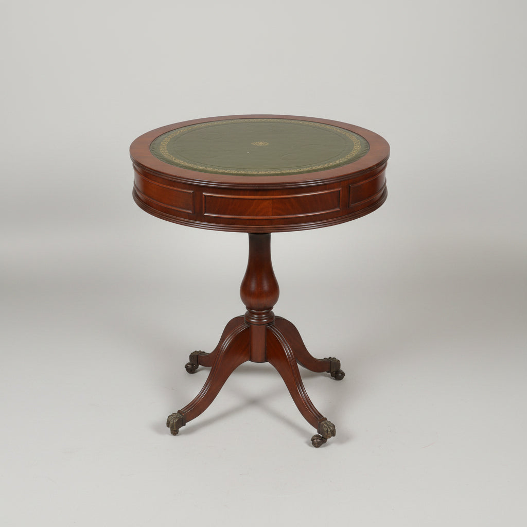 A round wooden pedestal table with a dark green and gold patterned top, photographed in a cluttered antique shop or furniture store.