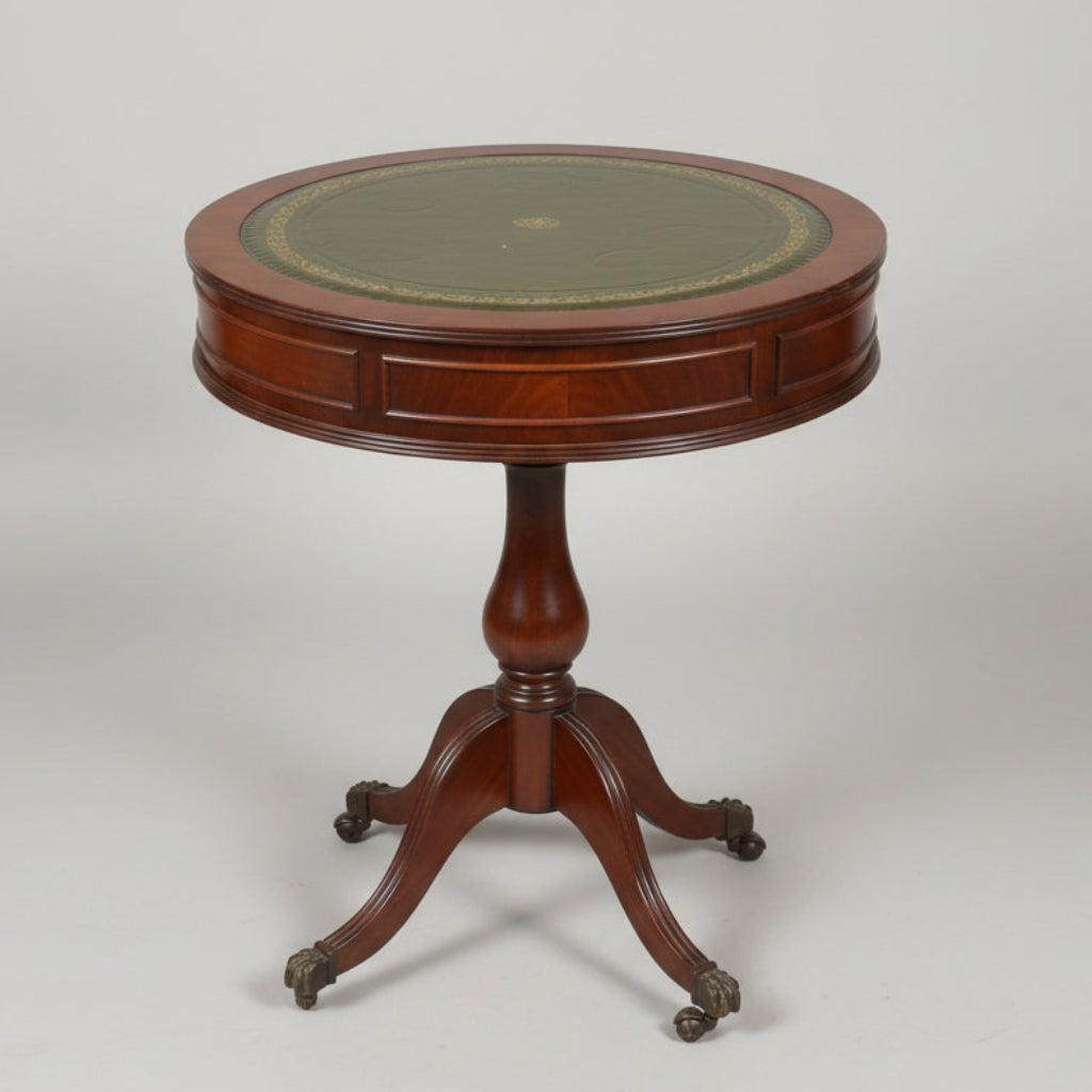A round wooden pedestal table with a dark green and gold patterned top, photographed in a cluttered antique shop or furniture store.