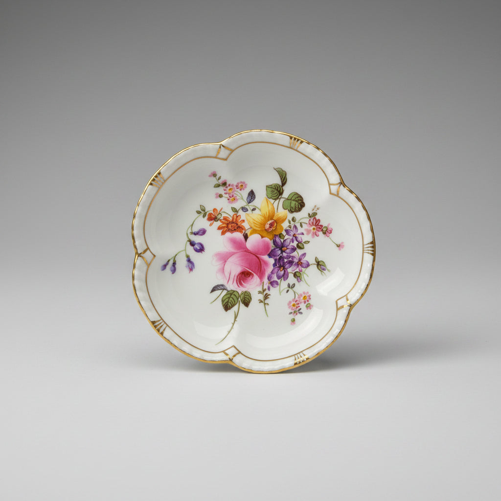 A scalloped white porcelain plate with gold trim and a colorful floral print, photographed from above on a dark wooden surface with a yellow patterned mat underneath.