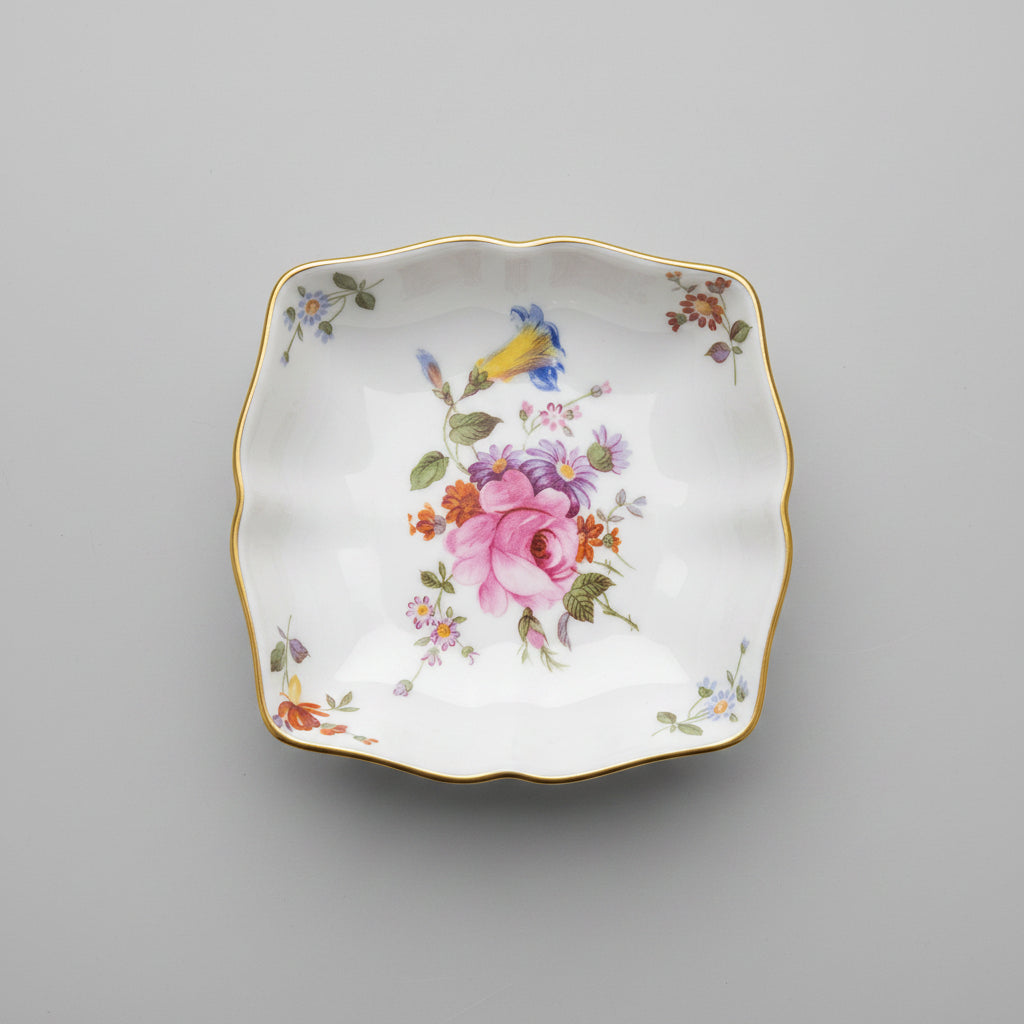 A vintage porcelain dish with a floral pattern, featuring a pink rose in the center and delicate gold trim, set against a dark wooden background with gold leaf detailing.