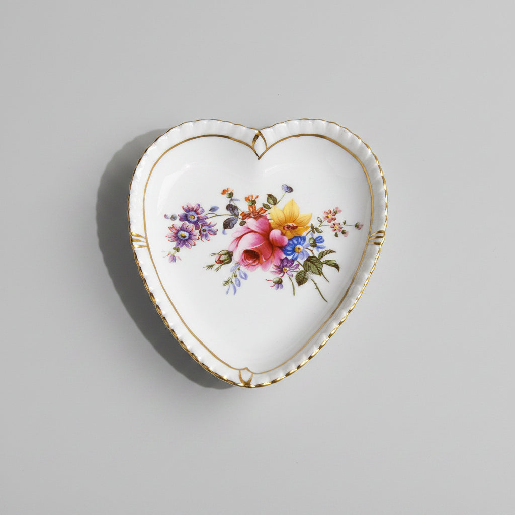 A heart-shaped white porcelain dish with gold trim and a colorful floral illustration in the center, placed on a carved wooden surface.