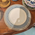 A ceramic dinner plate with a wide blue floral patterned border and a gold rim, layered over a white plate on a wooden table. Other dishes are partially visible around it.