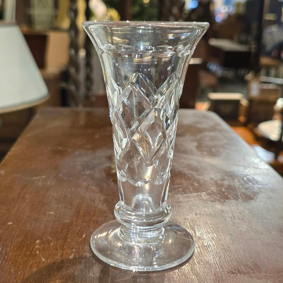 A clear cut-glass footed vase with a diamond pattern, standing on a dark wooden table in a cluttered antique shop.