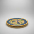A round ceramic serving platter with a blue and yellow border and green grape leaf pattern, displayed on a wooden table in a furniture store.