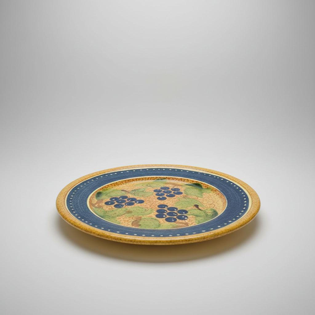 A round ceramic serving platter with a blue and yellow border and green grape leaf pattern, displayed on a wooden table in a furniture store.