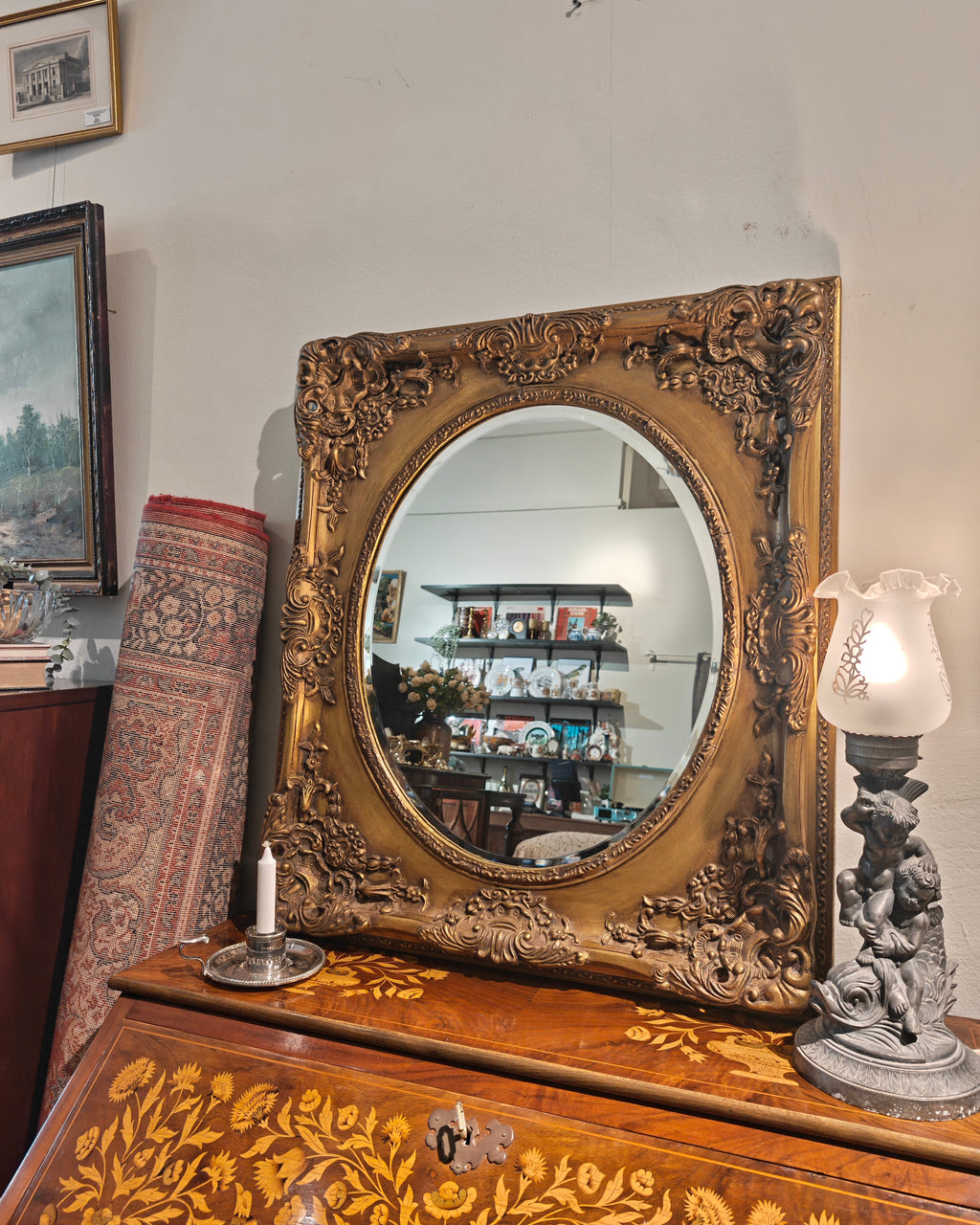 An ornate oval mirror with a carved gold frame resting on a decorated wooden chest of drawers in a vintage room. The mirror reflects shelves in the background, and a lit table lamp sits to the right.