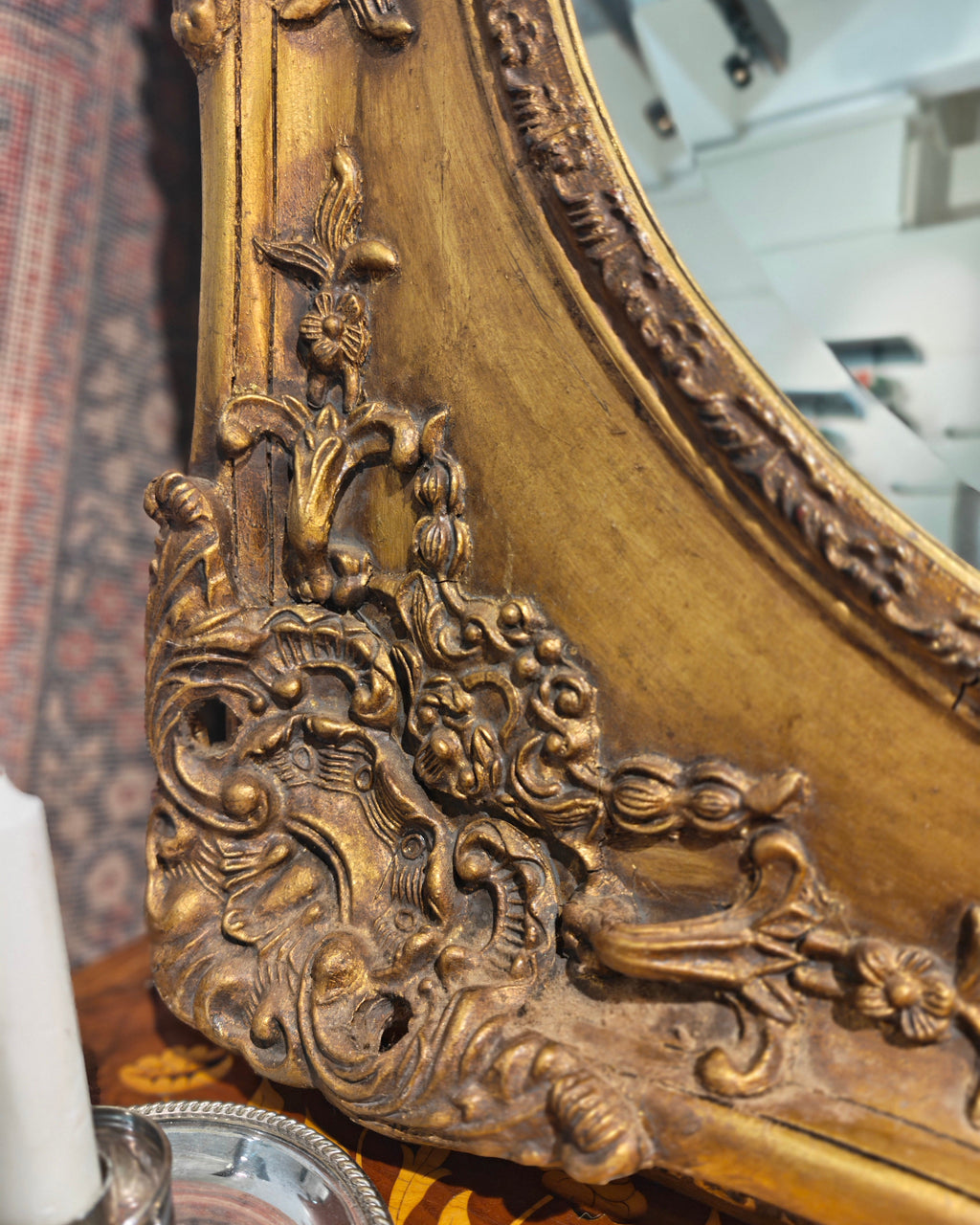 Close-up of an ornate gold mirror frame with intricate carved floral and scrollwork details, showing part of the reflective mirror surface.