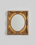 An ornate vintage-style oval mirror with a carved gold frame, displayed on a wooden cabinet in a home setting.