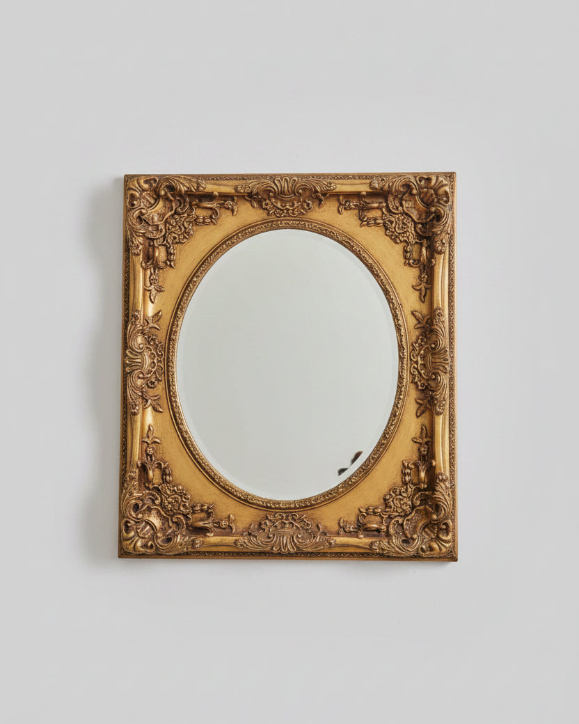 An ornate vintage-style oval mirror with a carved gold frame, displayed on a wooden cabinet in a home setting.