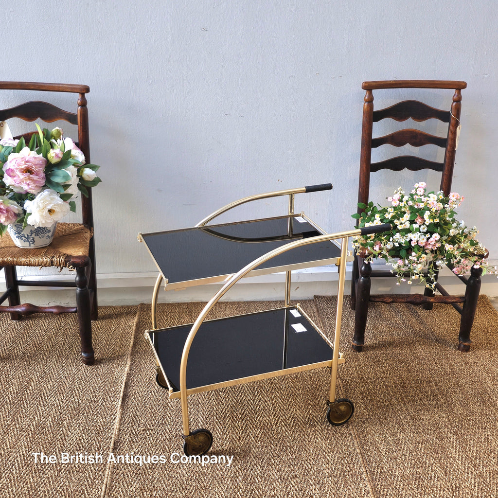 Vintage-style gold and black serving cart with two wooden chairs and floral arrangements on a textured floor.