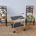 Vintage-style gold and black serving cart with two wooden chairs and floral arrangements on a textured floor.
