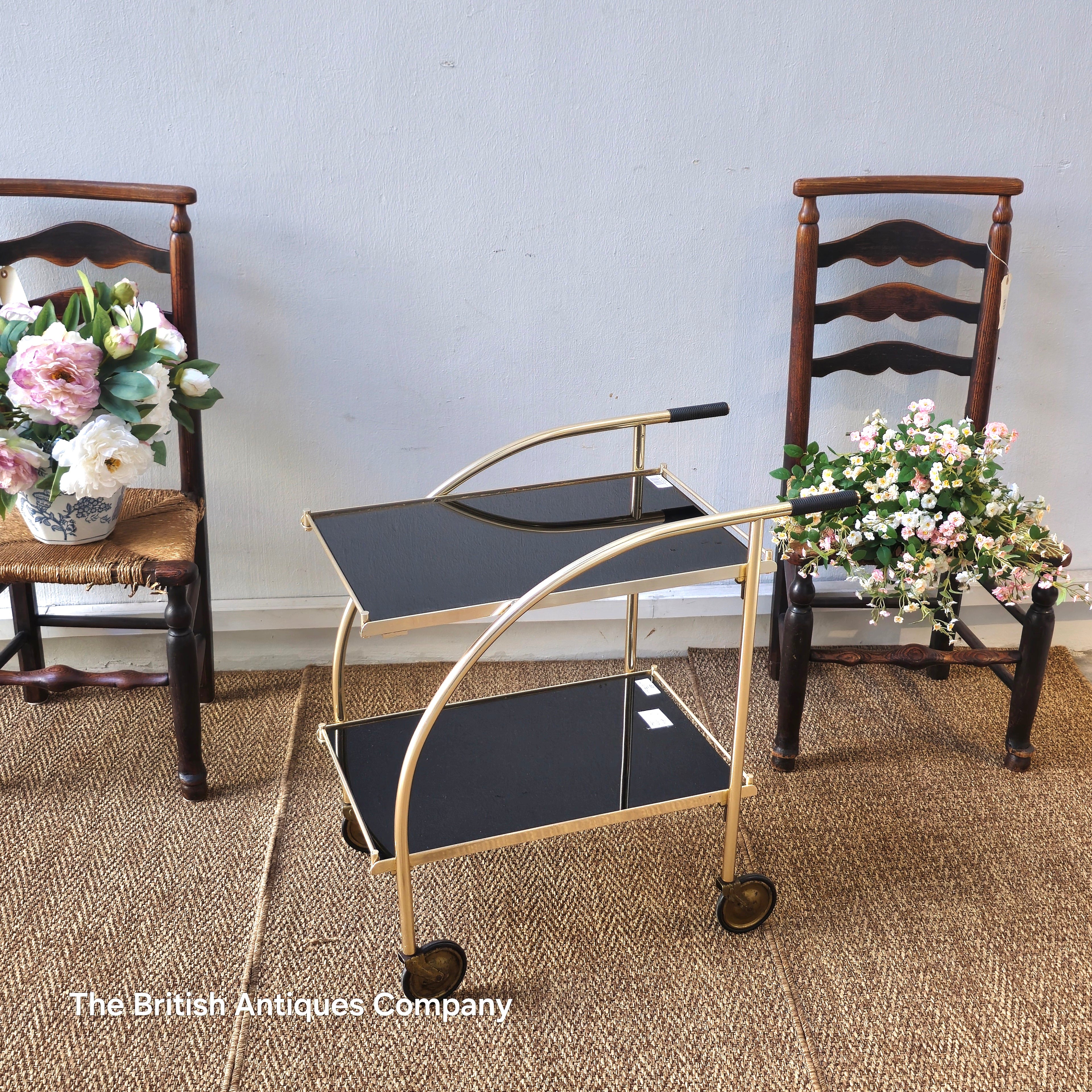 Vintage-style gold and black serving cart with two wooden chairs and floral arrangements on a textured floor.