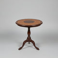 A round wooden side table with a dark metal pedestal base, featuring a light tan woven cane top with a dark brown circular center medallion, set on a distressed red and green floor in front of red stairs.