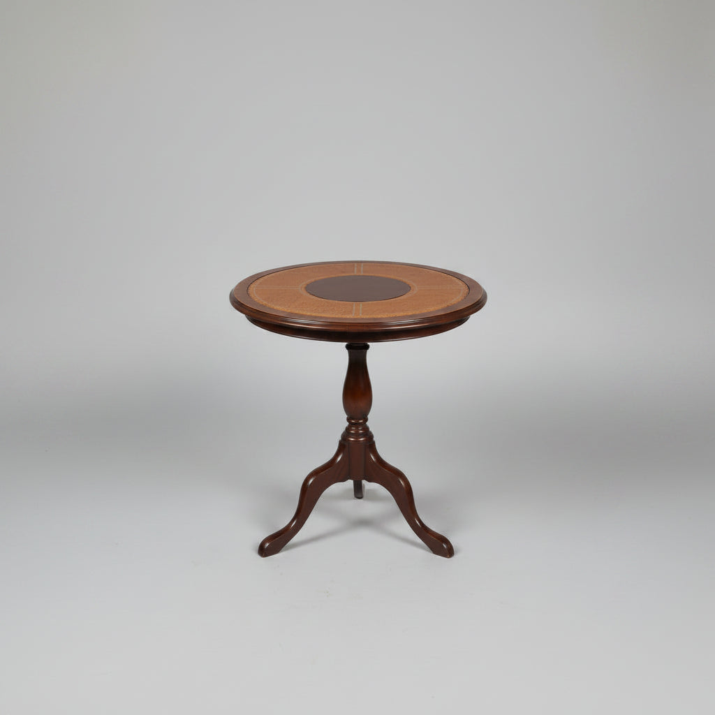 A round wooden side table with a dark metal pedestal base, featuring a light tan woven cane top with a dark brown circular center medallion, set on a distressed red and green floor in front of red stairs.