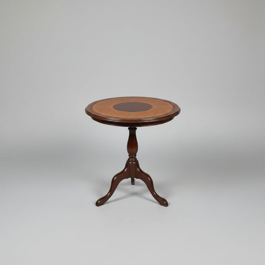 A round wooden side table with a dark metal pedestal base, featuring a light tan woven cane top with a dark brown circular center medallion, set on a distressed red and green floor in front of red stairs.