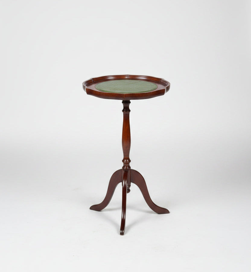 A small round wooden side table with a green top and three legs, photographed in a warehouse or showroom setting on a worn red-brown floor.