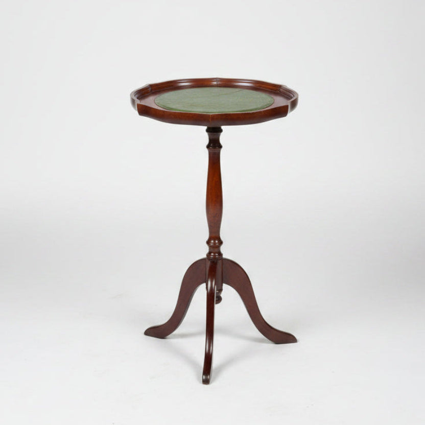 A small round wooden side table with a green top and three legs, photographed in a warehouse or showroom setting on a worn red-brown floor.