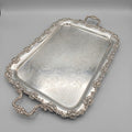 An ornate rectangular silver serving tray with decorative handles and a raised floral border, photographed in a real indoor room with carpet, furniture, and framed items visible in the background.