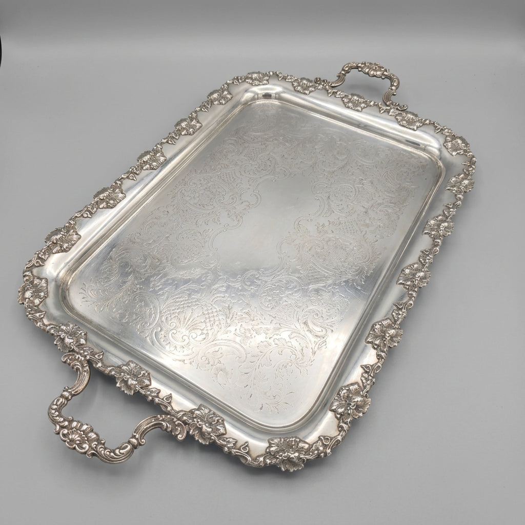 An ornate rectangular silver serving tray with decorative handles and a raised floral border, photographed in a real indoor room with carpet, furniture, and framed items visible in the background.