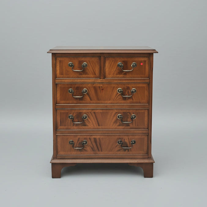 A vintage wooden chest of drawers with four drawers and ornate dark metal handles, styled in a cluttered antique shop next to a purple velvet armchair and a glass display cabinet.