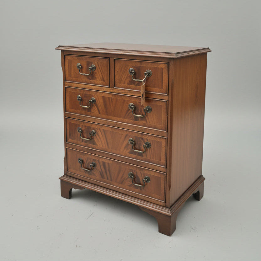 A vintage wooden chest of drawers with five drawers and ornate dark metal handles, situated in a cluttered antique shop. Decorative ceramic vases and a lamp sit on top of the chest.