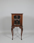 An antique dark wood cabinet with glass doors stands in a shop interior next to a woven-back chair with Union Jack flags. The cabinet is decorated with a brass vase of flowers and candlesticks on top.