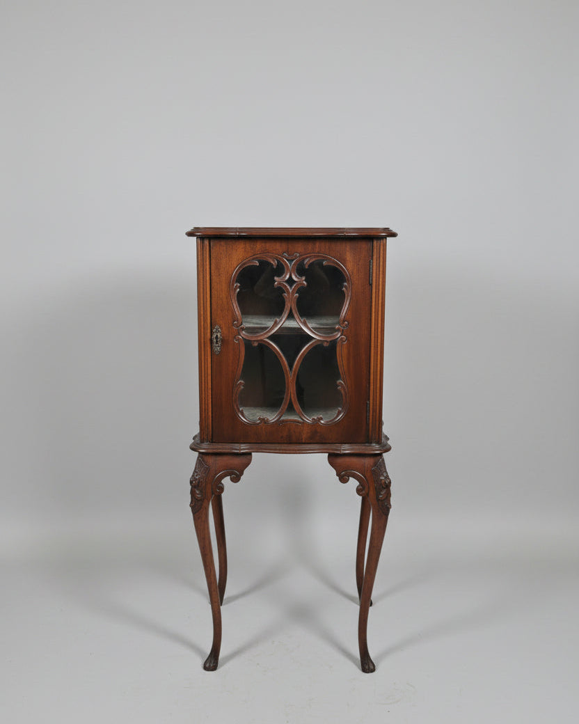 An antique dark wood cabinet with glass doors stands in a shop interior next to a woven-back chair with Union Jack flags. The cabinet is decorated with a brass vase of flowers and candlesticks on top.