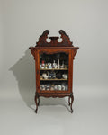 An ornate dark wood cabinet with glass doors and carved details, filled with teapots and ceramic items, photographed in a furniture showroom.