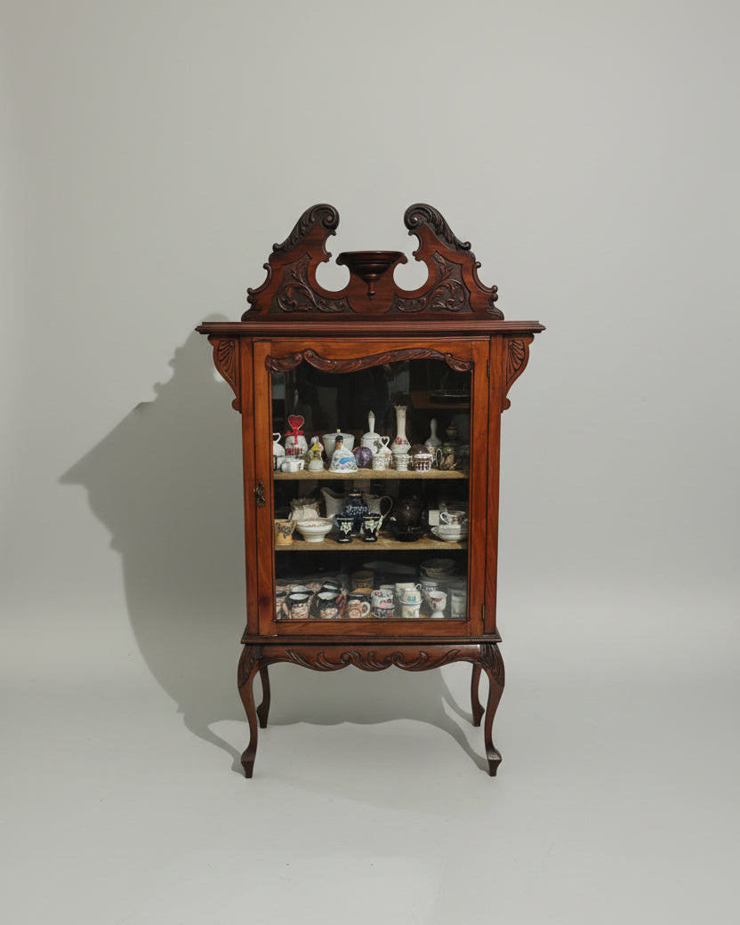 An ornate dark wood cabinet with glass doors and carved details, filled with teapots and ceramic items, photographed in a furniture showroom.