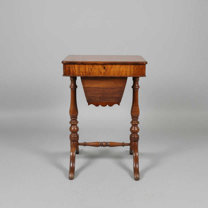 An antique wooden footstool or small side table with turned legs and a polished wood top, photographed in a furniture showroom with other wooden pieces in the background.