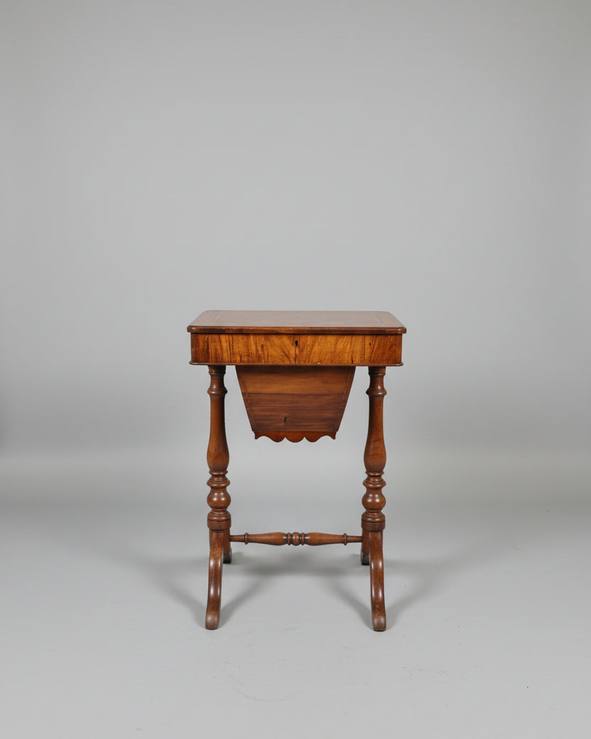 An antique wooden footstool or small side table with turned legs and a polished wood top, photographed in a furniture showroom with other wooden pieces in the background.