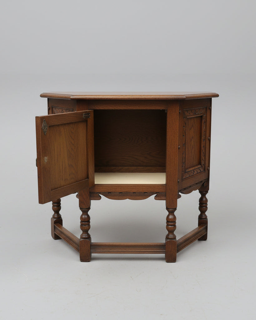 A dark brown wooden side table or cabinet with turned legs and an open door showing an interior shelf, photographed in a furniture store or warehouse setting.